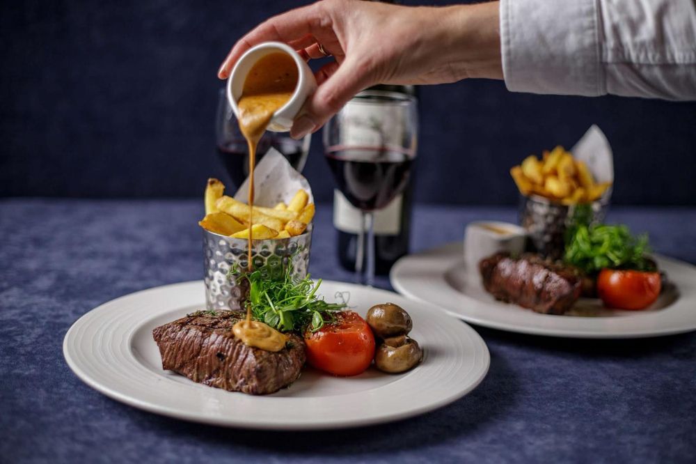 Two steak plates on navy linen, peppercorn sauce being poured over a cup of fries