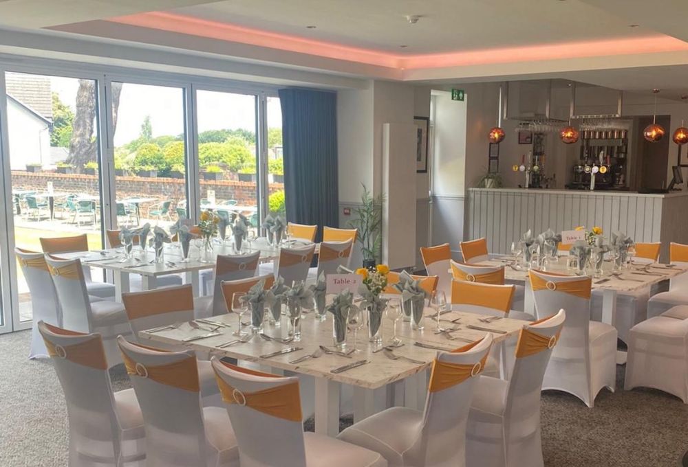 The snug at The Balgarth Pines laid out for a party, long tables with white chair covers and orange sashes, bifold doors onto the terrace and the bar at the far end under copper pendant lights