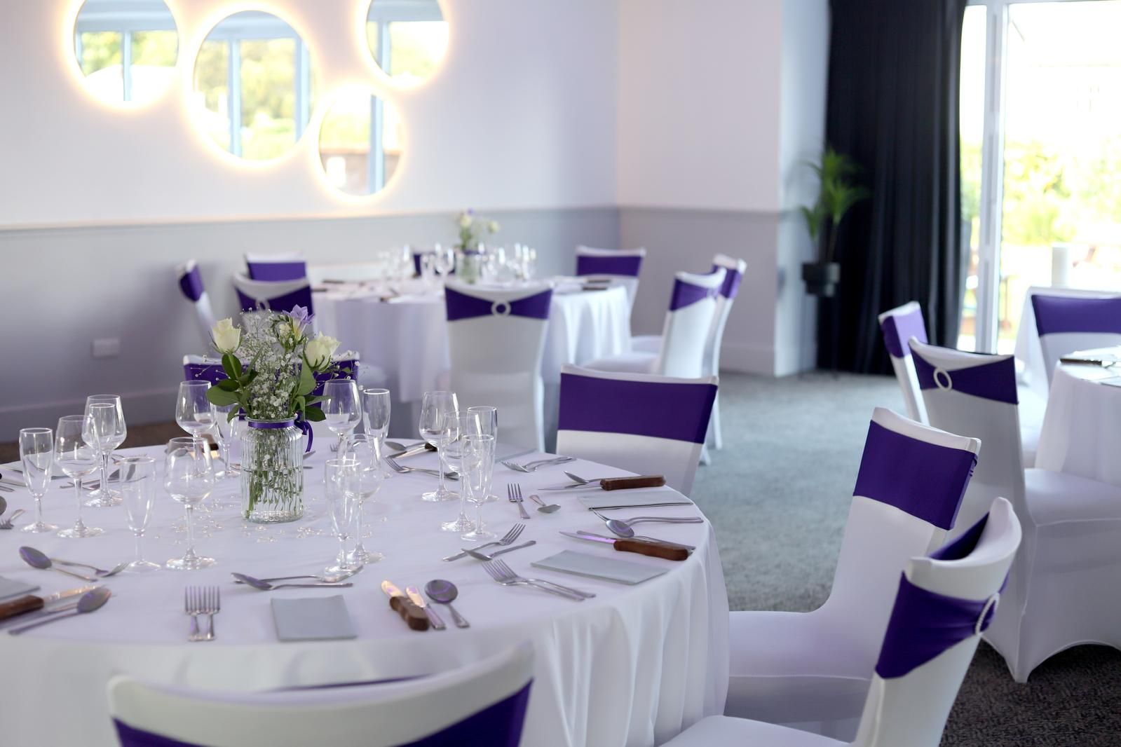 A round table at The Balgarth Pines dressed in white linen with purple sashes, illuminated circular mirrors on the wall behind and the garden through the glazing